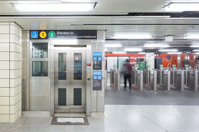Elevator in Bloor-Yonge Station to Line 1 Northbound, and Like 2 Eastbound and Westbound. The stainless steel elevator is to the left with 6 Presto turnstiles to the left. One person is going through the turnstile. The wall is orange tile in the background.