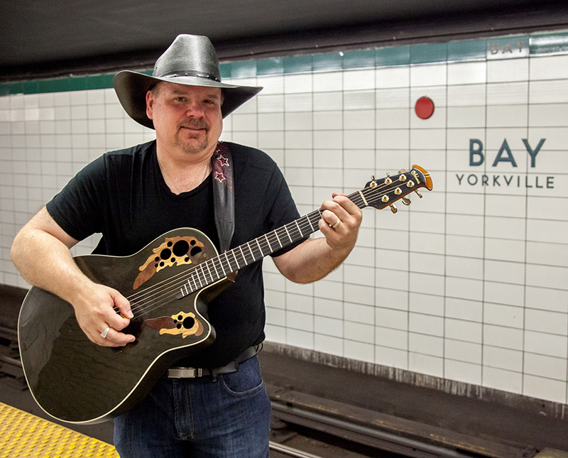 Blair Franklin smiles and wear a cowboy hat, in front of a subway station platform.