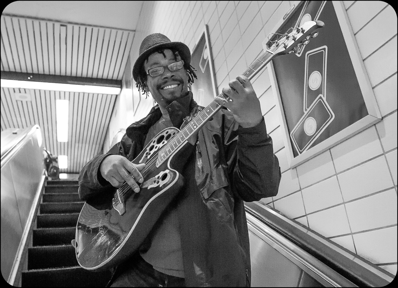 Adam smiles with a guitar in hand, infront of a subway station stairwell.
