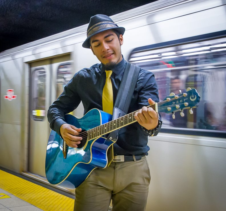 Morad Saad Guzman playing a blue guitar while a subway passes by in the background.