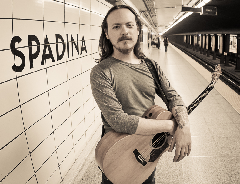 Rob London stands on a subway platform with a guitar hanging around his neck.