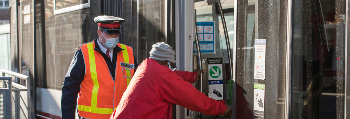 A TTC bus operator wearing a face mask assists a customer, who is also wearing a face mask, board the bus on a sunny day.