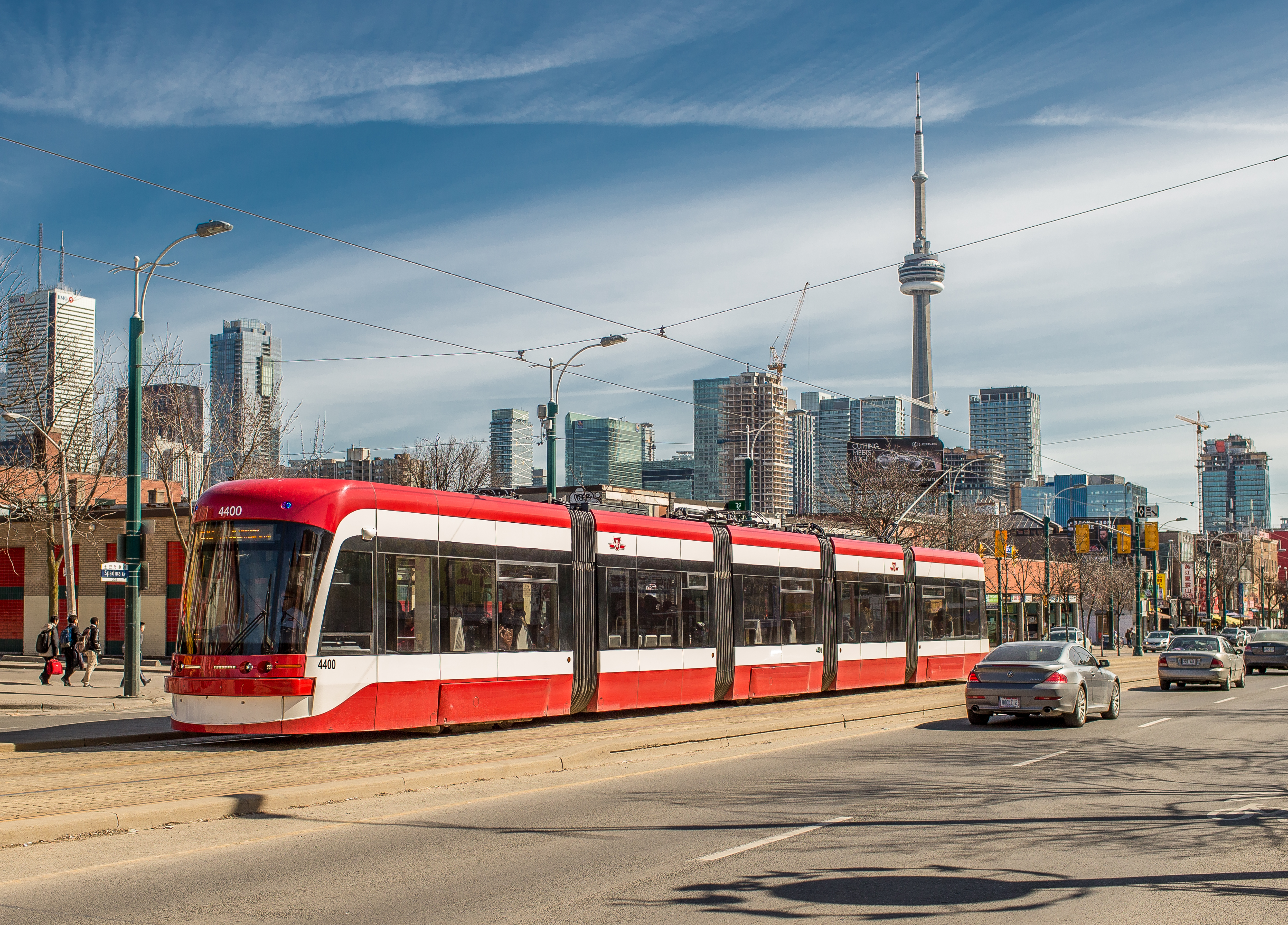 TTC streetcar traveling along a city street with cars and pedestrians nearby, set against the Toronto skyline with the CN Tower rising in the background under a blue, partly cloudy sky.