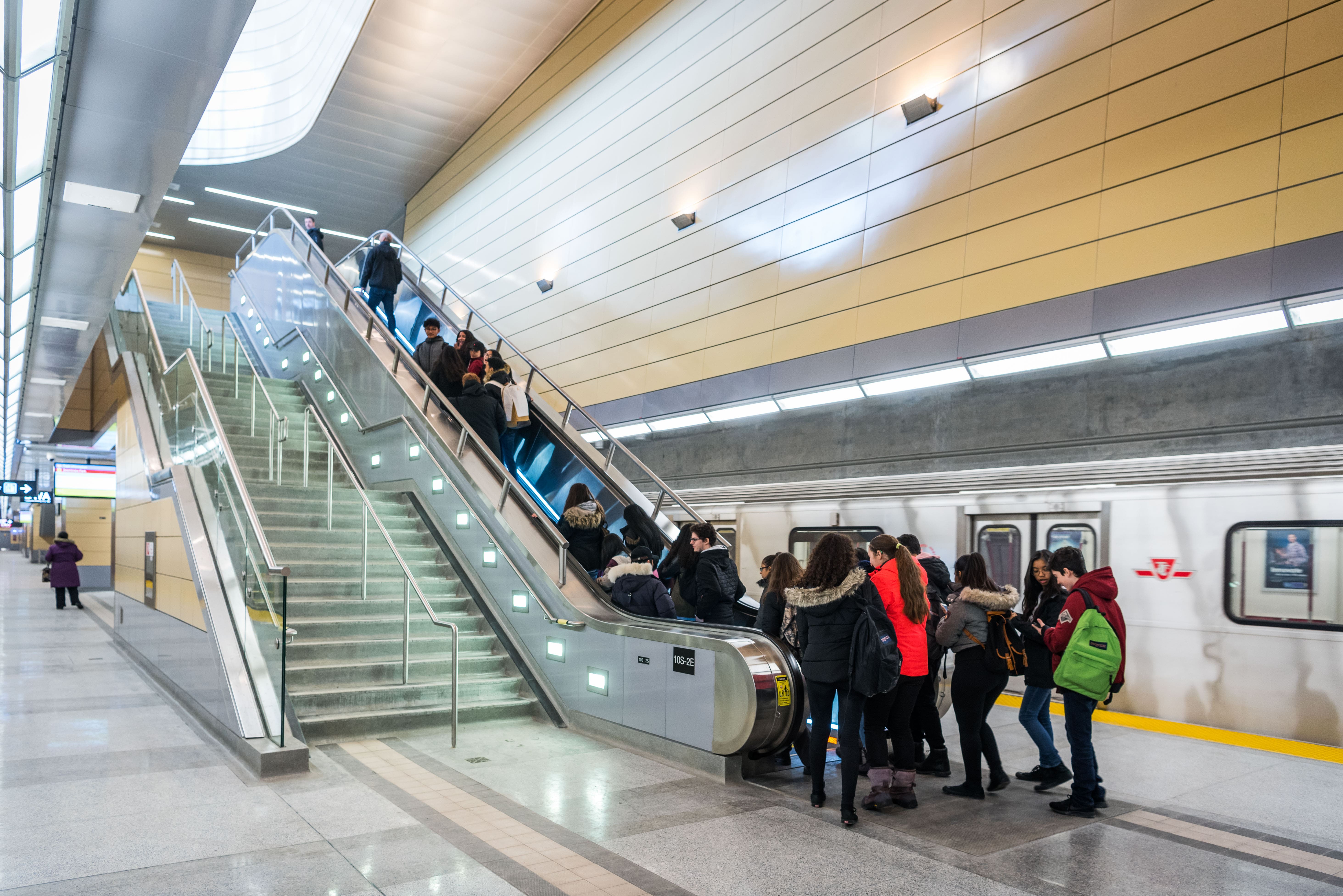 Group of students boarding an escalator on a subway station platform with a TTC subway train in the background.