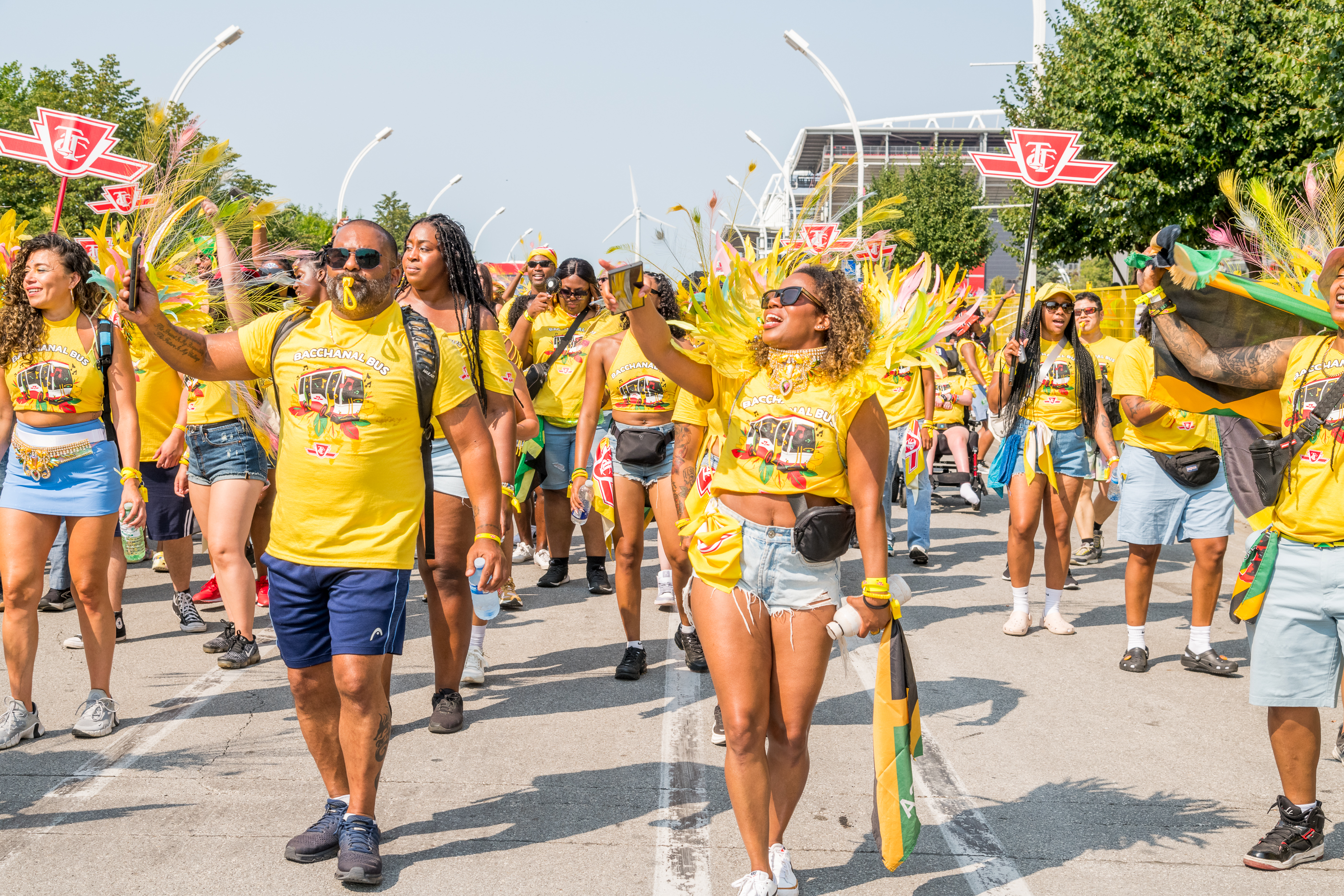 Large group of people wearing matching yellow shirts walking down a street at the Toronto Caribbean Carnival Grand Parade. Some people are holding red and white TTC signs.