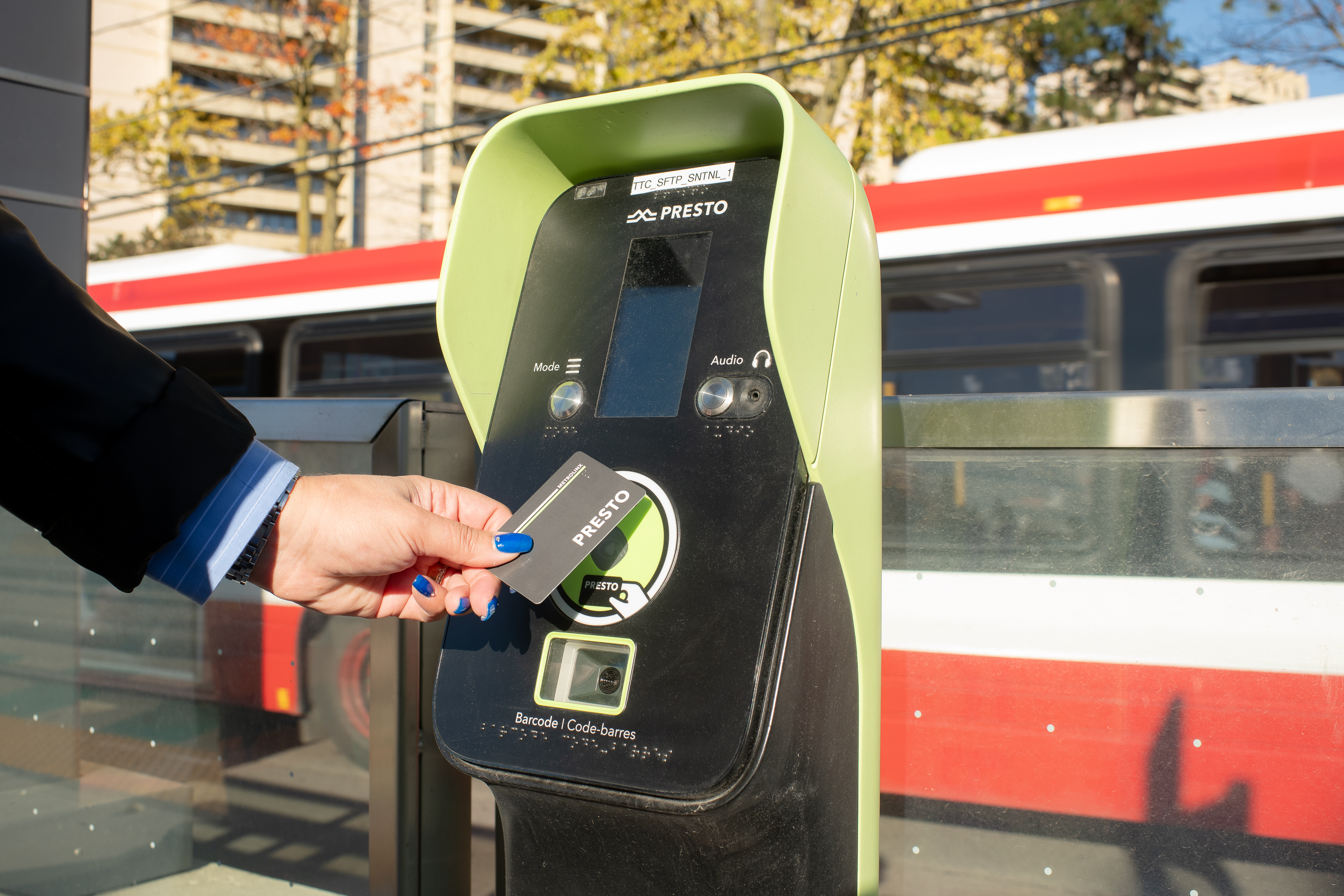 A hand tapping a PRESTO card on a PRESTO reader with a red and white TTC bus in the background.