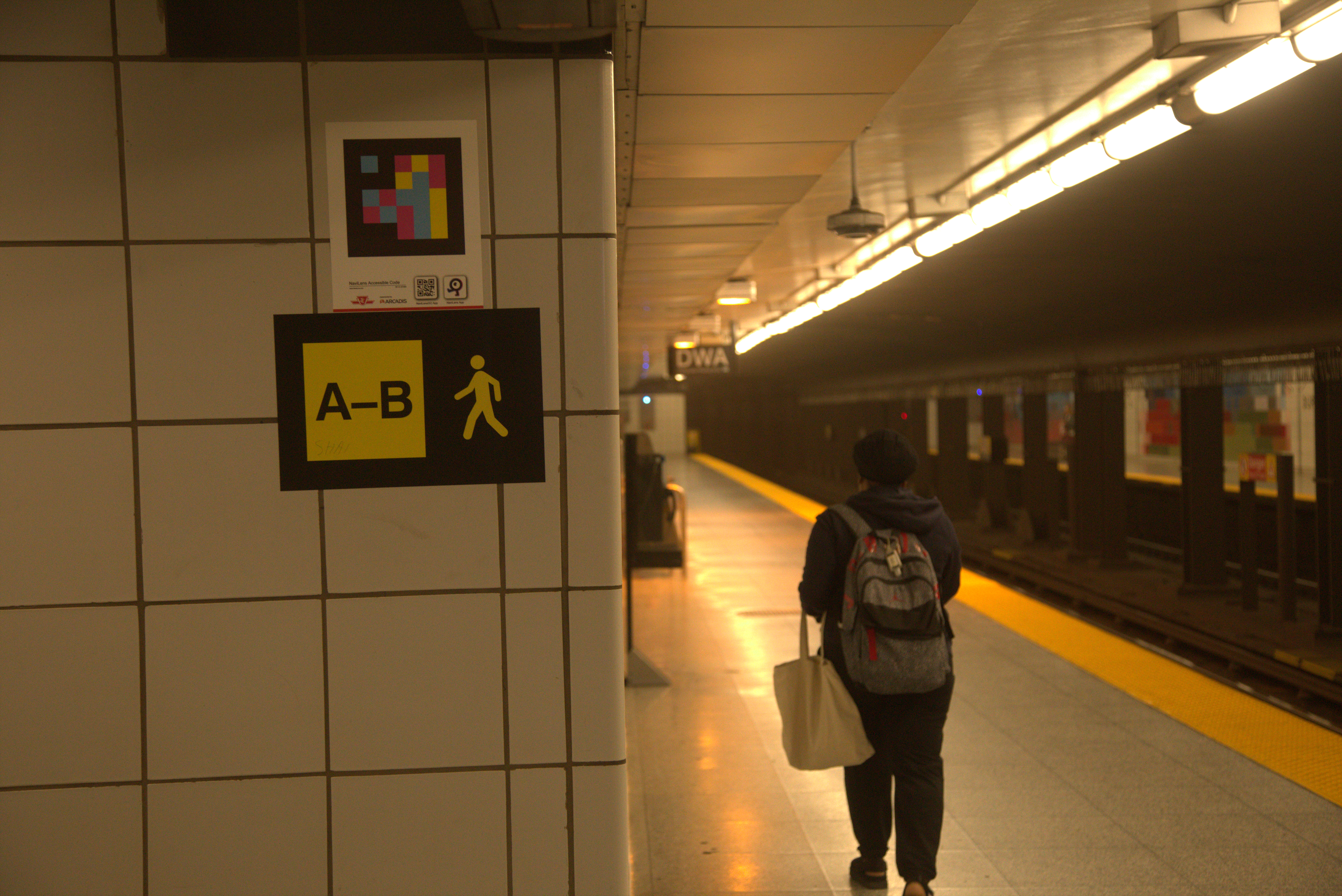 Subway station wall with a colourful NaviLens code and a black and yellow sign with the letters A-B and symbol of a person walking. A person is walking on the platform in the background.