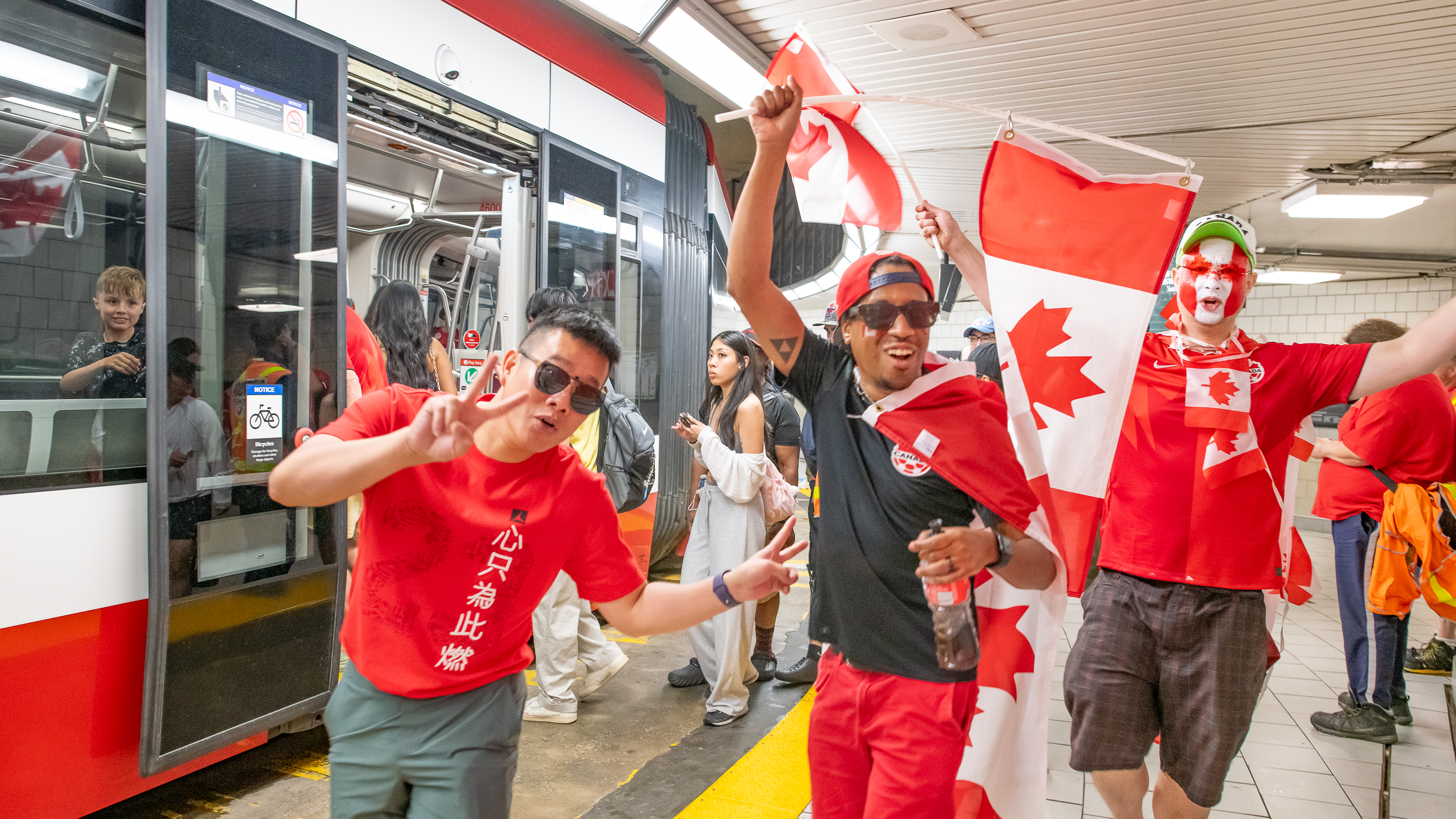 Three soccer fans wearing red Canada-themed clothes and holding Canadian flags in front of a TTC streetcar