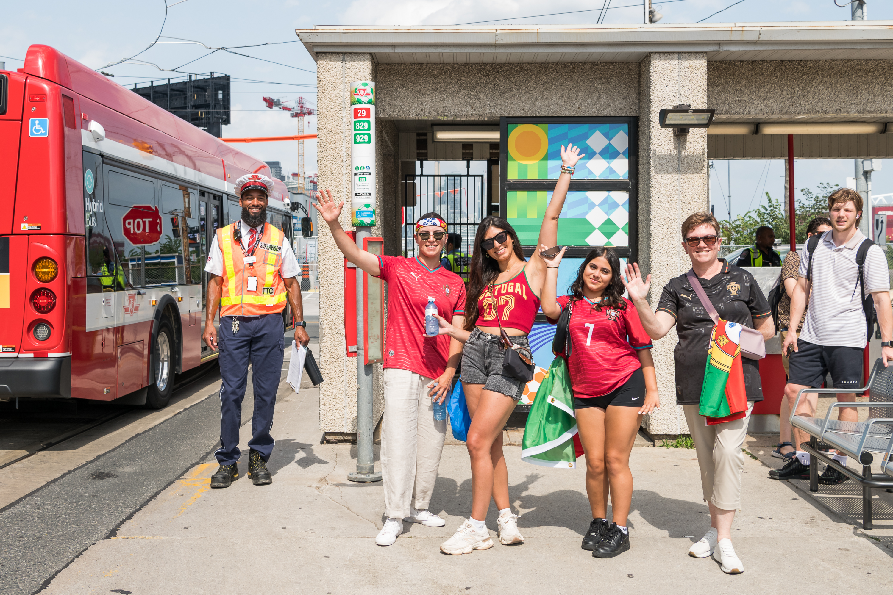 Portugal fans in Portugal themed outfits and a TTC operator posing for a photo at a bus stop. 