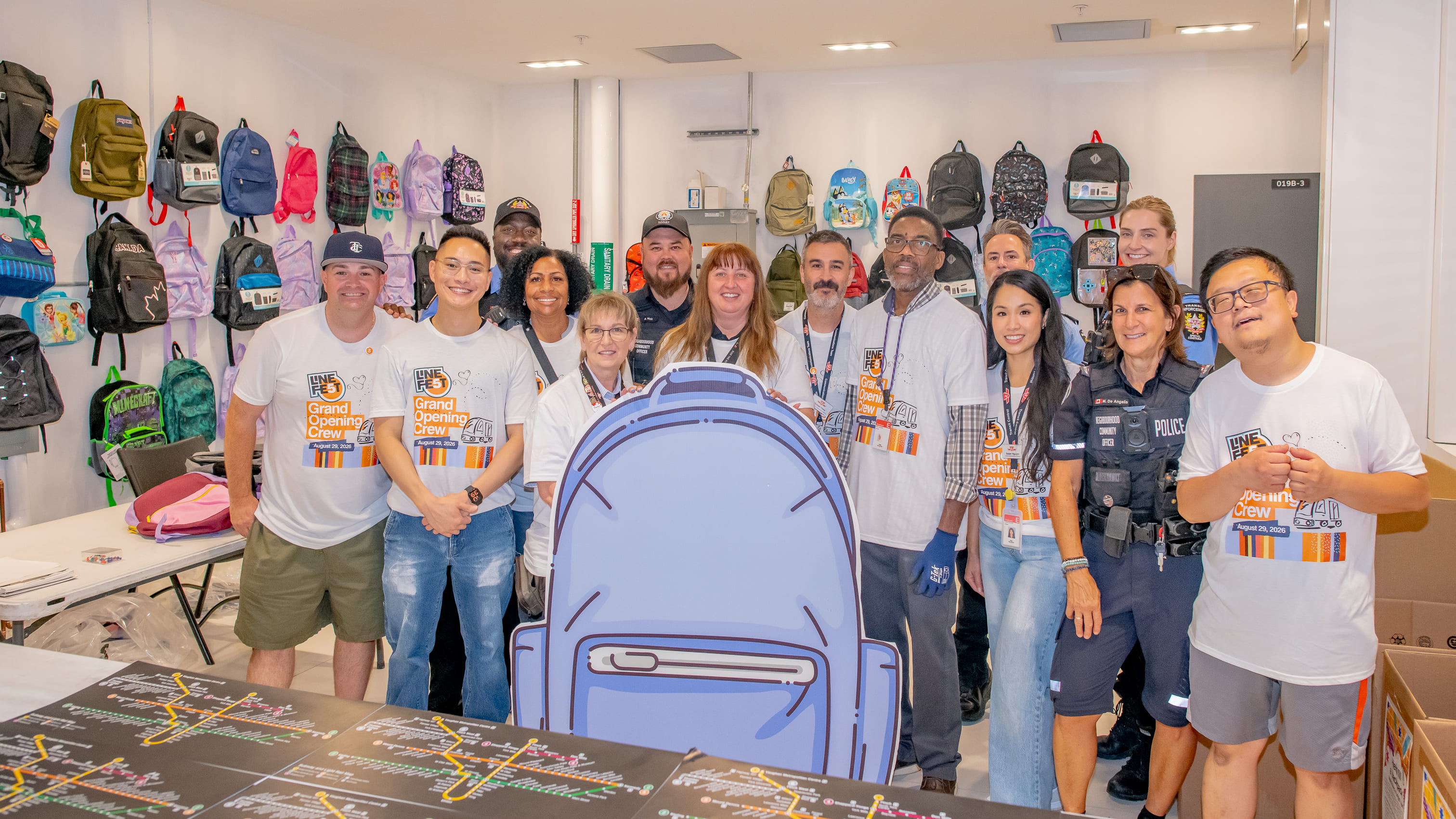 TTC staff pose for a photo around a backpack cutout with backpacks in the background, to celebrate the final day of the TTC Backpack Drive