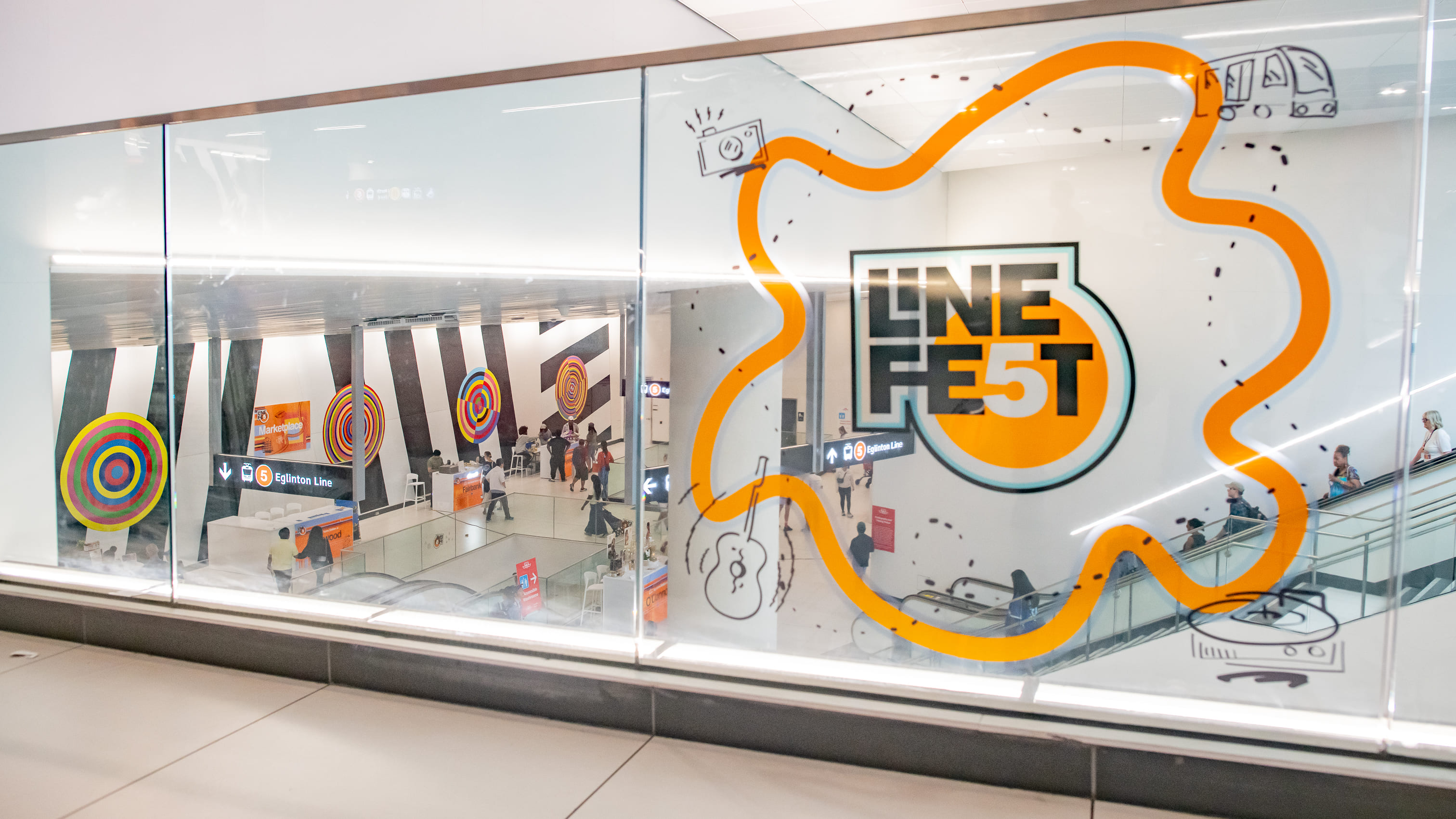 Decorative Line 5 Fest graphic on a glass wall, with the Eglinton Station concourse and celebration setup in the background