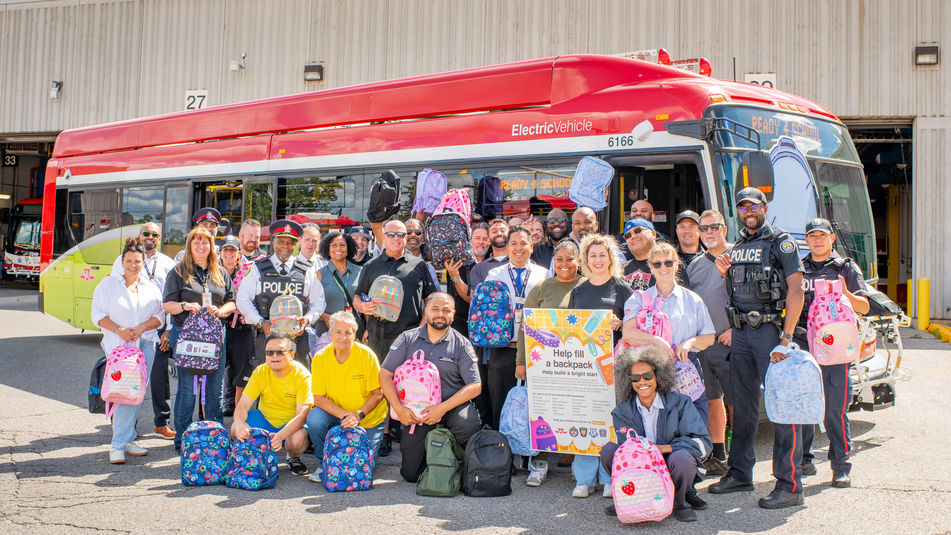 Group photo of TTC staff, special constables, Toronto Police, and other partners, holding donated backpacks in front of a parked TTC Backpack Drive bus