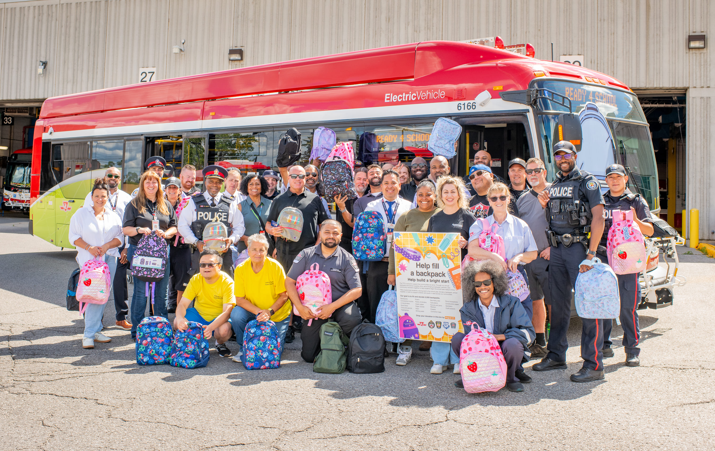 Group photo of TTC staff, special constables, Toronto Police, and other partners, holding donated backpacks in front of a parked TTC Backpack Drive bus
