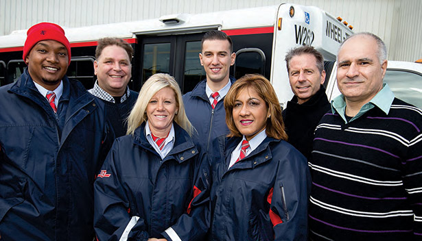 Several employees smile at the camera, in front of a Wheel-trans vehicle.