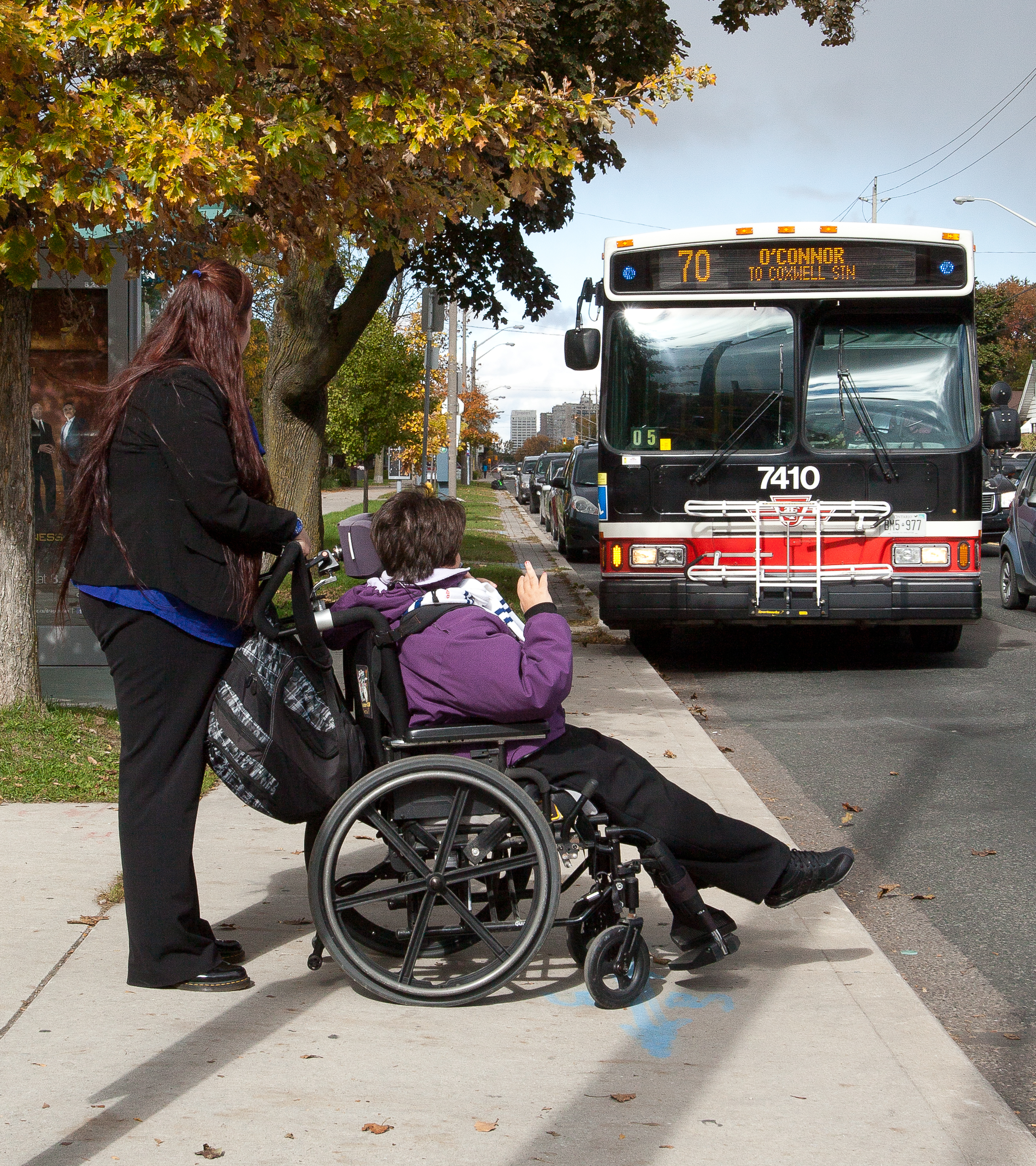 Two women, one sitting in a wheel-chair and the other standing behind and holding the wheel-chair, are on a sidewalk looking at an approaching TTC bus. 